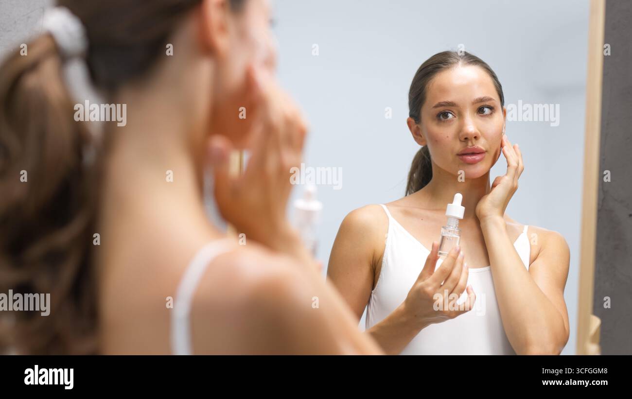 Una giovane donna sta applicando un siero per la cura della pelle mentre guarda in uno specchio, evidenziando la sua pelle sana e il suo regime di bellezza, circondato da un bagno rilassante Foto Stock