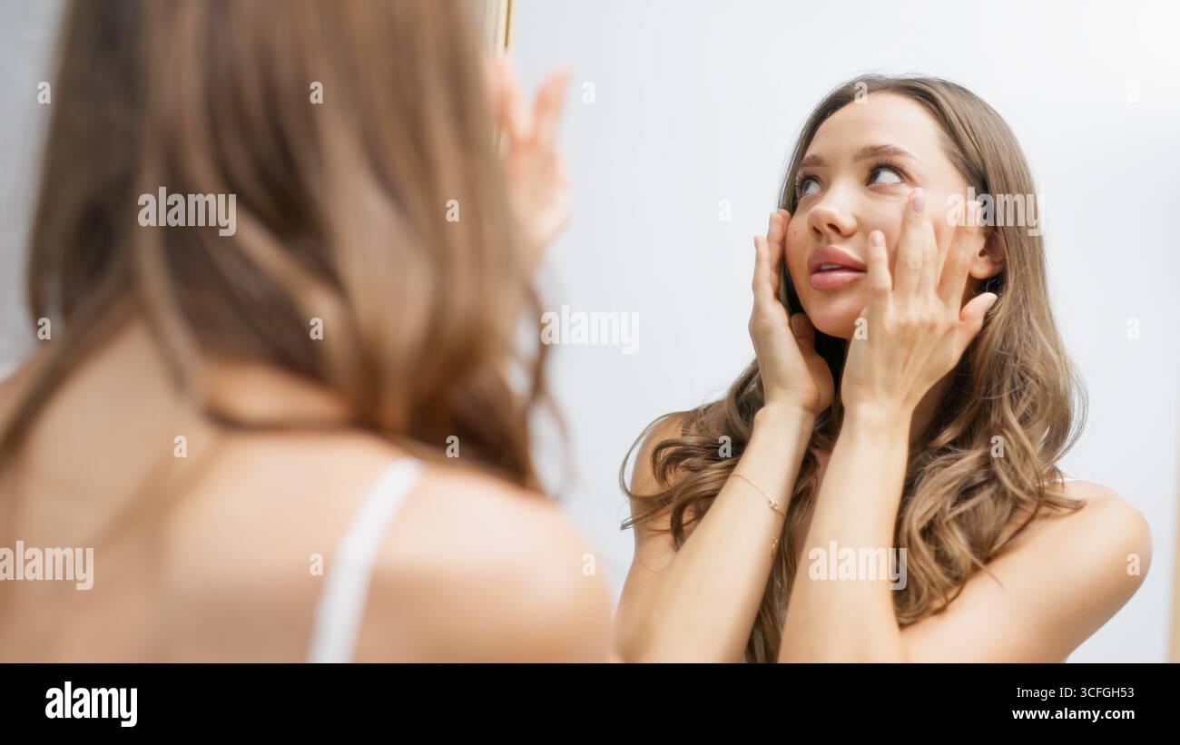 La giovane donna con lunghi capelli castani sta applicando delicatamente un prodotto per la cura della pelle sul viso mentre guarda allo specchio, sottolineando il suo impegno per una guarigione Foto Stock