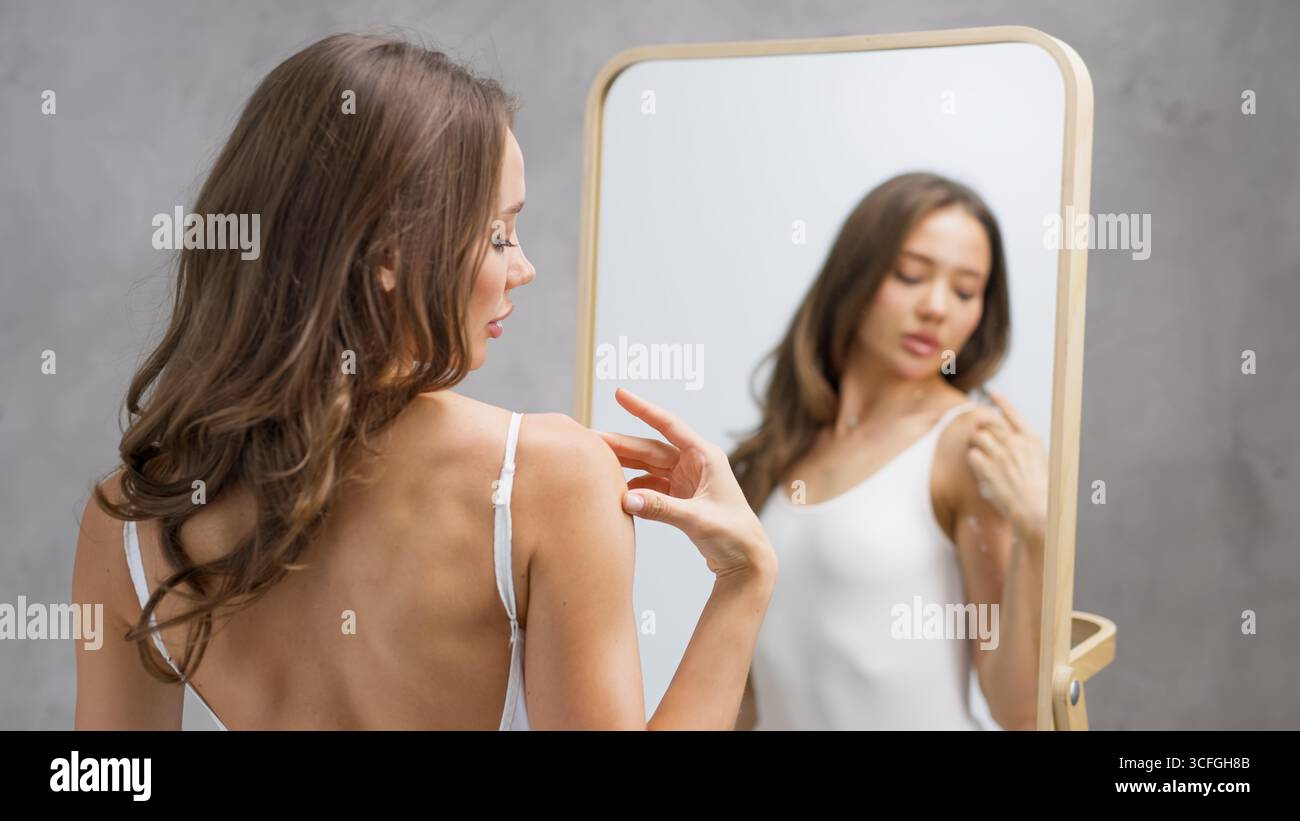 Donna con lunghi capelli castani sta applicando un prodotto per la cura della pelle mentre si guarda in uno specchio, evidenziando la sua pelle sana e la routine di benessere in un indaco calmo Foto Stock