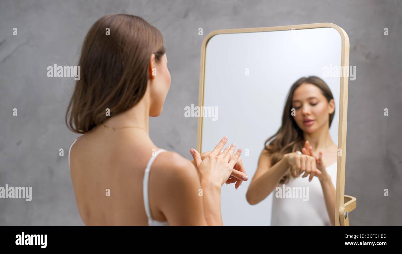 La giovane donna sta applicando un prodotto per la cura della pelle sulle mani mentre guarda in uno specchio, evidenziando la sua pelle sana e la routine di benessere, circondato da Foto Stock