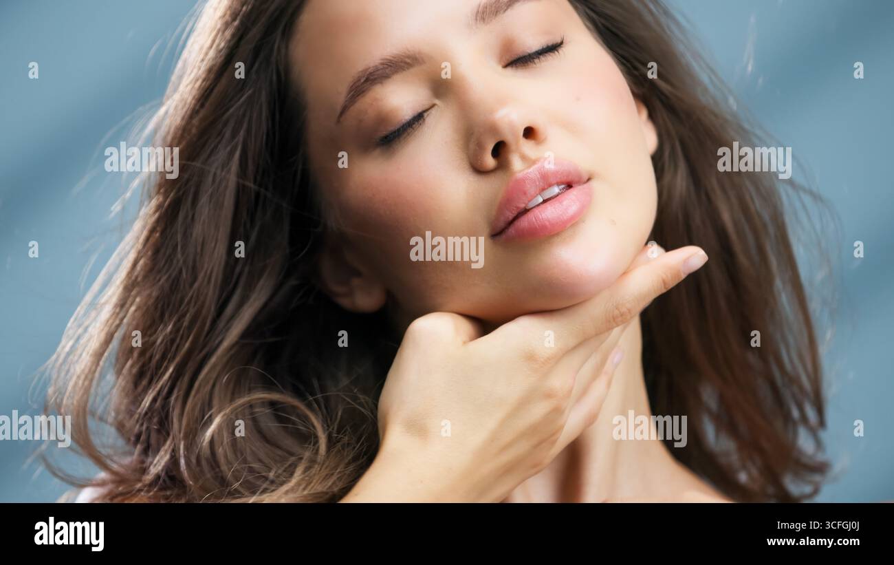 La donna con una pelle impeccabile tocca delicatamente il viso, evidenziando la sua colorazione radiosa e la sua bellezza naturale, circondata da una luce soffusa che migliora Foto Stock