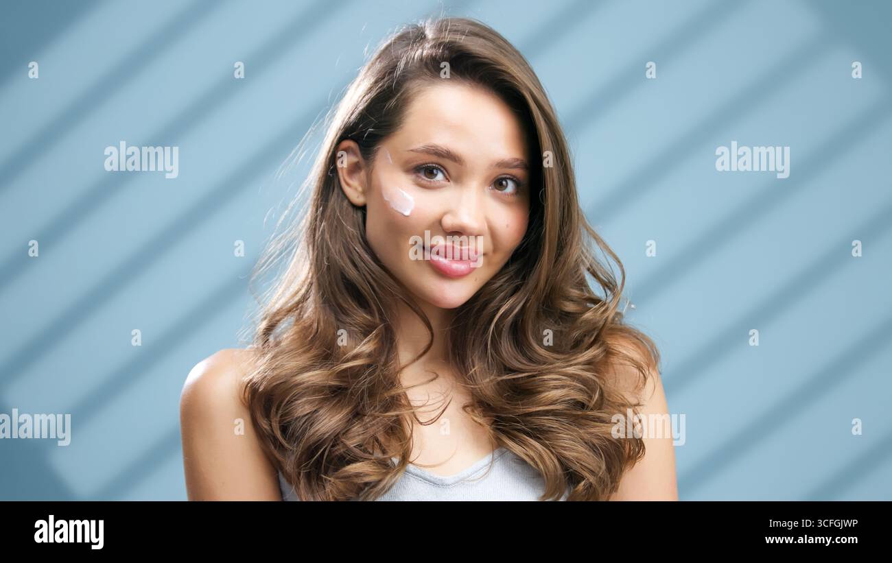 La giovane donna con la pelle luminosa sta applicando un idratante sulla guancia, evidenziando la sua colorazione radiosa, circondata da una luce soffusa e da un bac sfocato Foto Stock