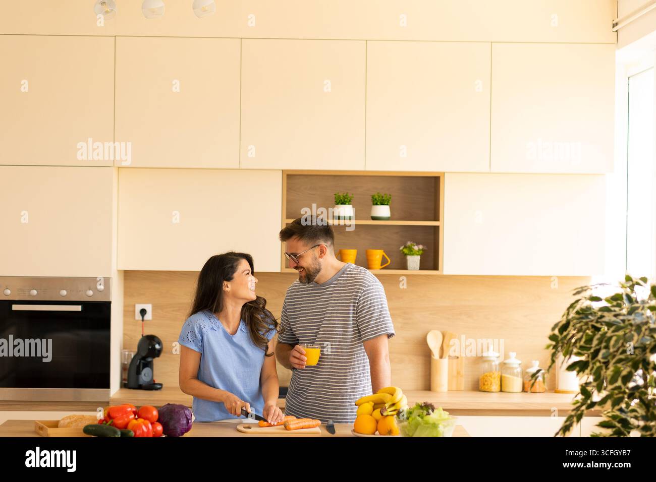 All'interno di una cucina spaziosa e contemporanea, due amici condividono un momento di gioia mentre tritano verdure e gusteranno bevande. Vengono preparati ingredienti freschi Foto Stock
