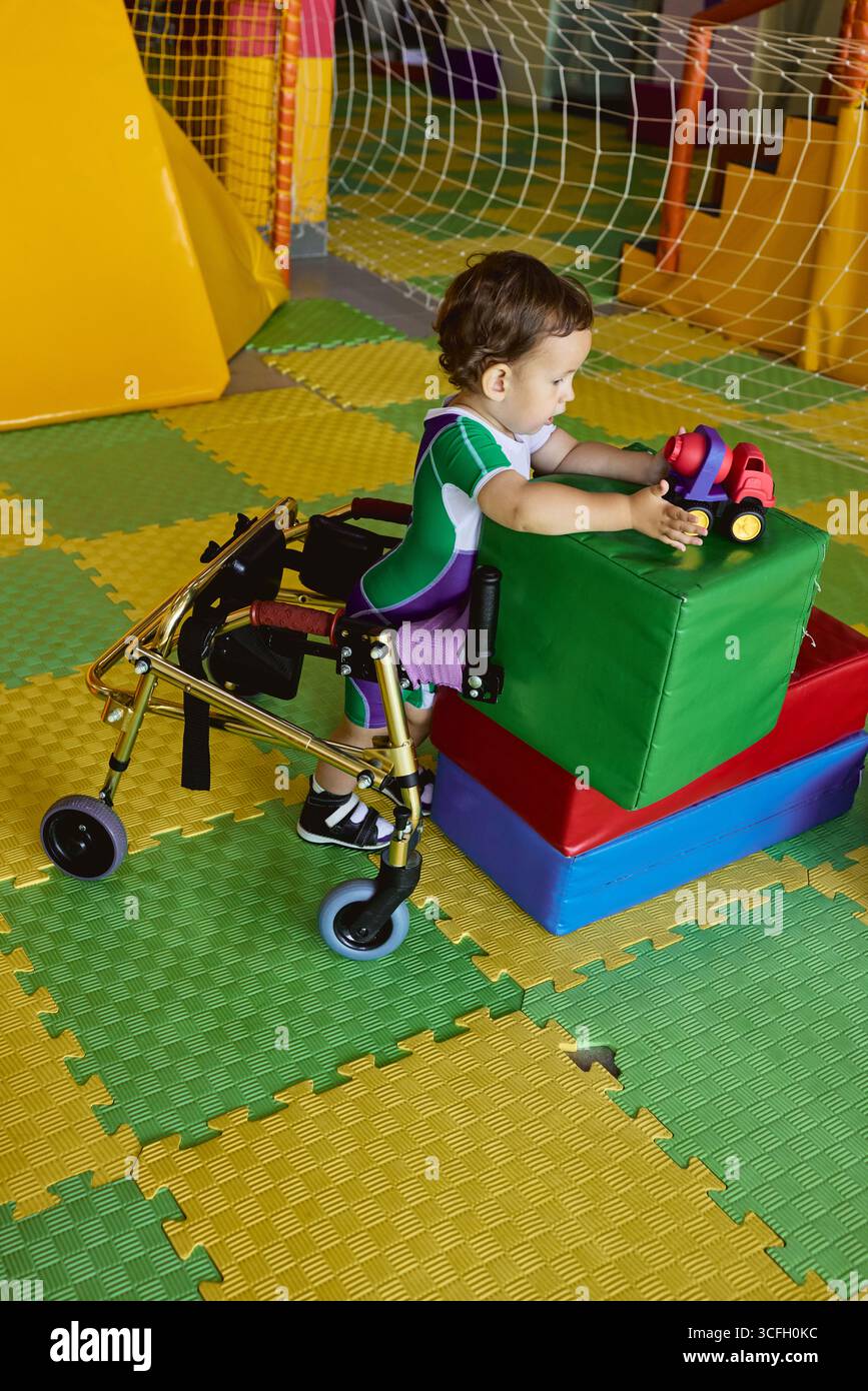 Il bambino gioioso usa un camminatore in fisioterapia, circondato da colorate attrezzature di riabilitazione. Recupero dei figli Foto Stock