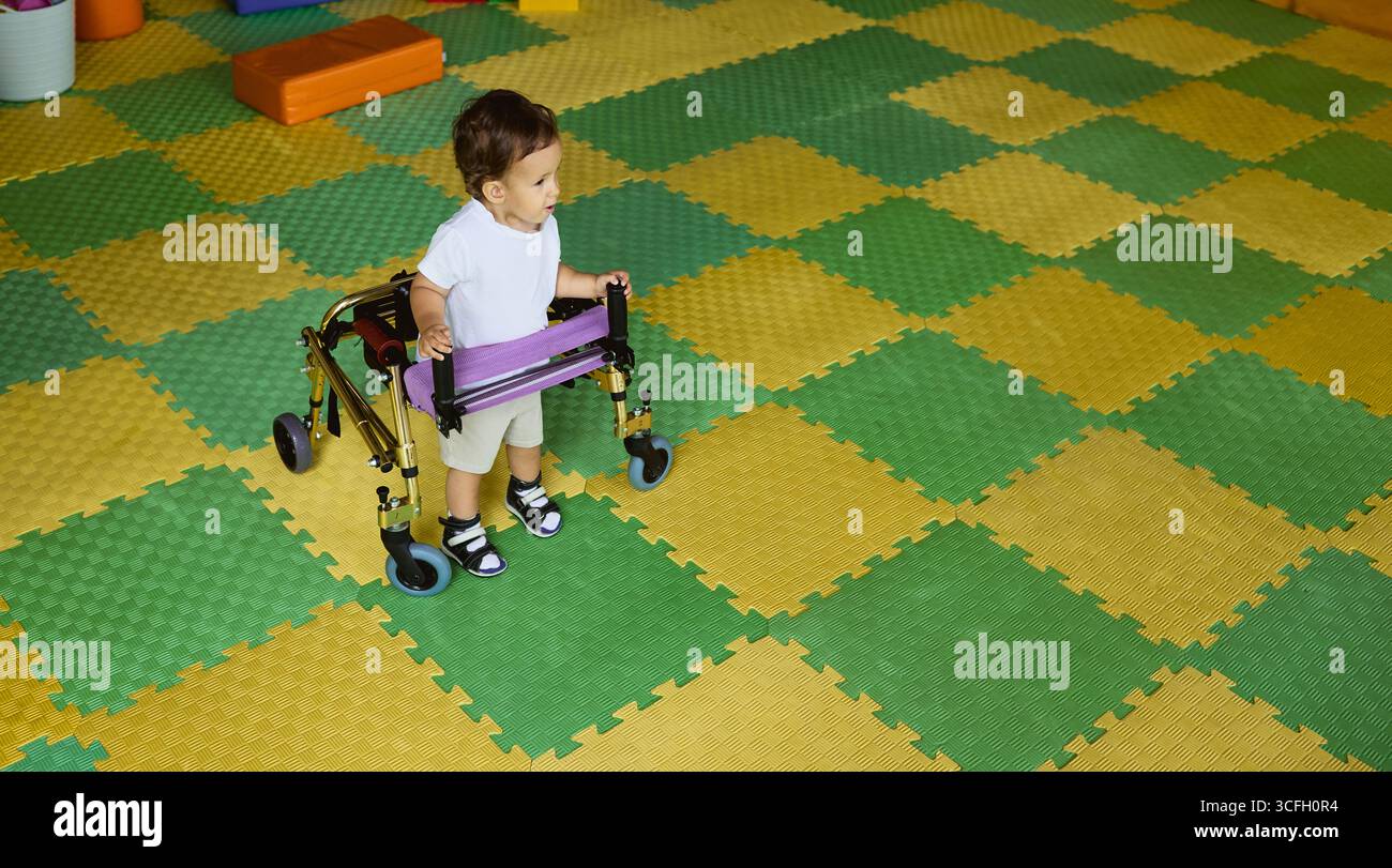 Riabilitazione bambini. Il bambino pratica a camminare con un robusto camminatore, circondato da tappeti vibranti che incoraggiano il movimento e il gioco Foto Stock