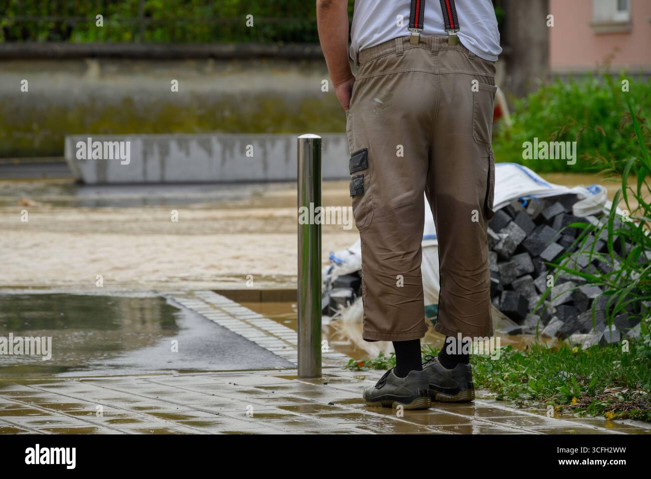 Un uomo osserva il terreno bagnato accanto ai materiali da costruzione dopo le forti piogge in un'area residenziale, mostrando l'impatto delle inondazioni. Foto Stock