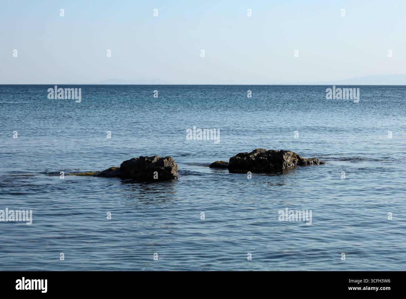 Tranquillo scenario costiero caratterizzato da due rocce scure che emergono dal mare calmo, con un orizzonte pulito e un cielo azzurro morbido sullo sfondo. Minimale e pe Foto Stock