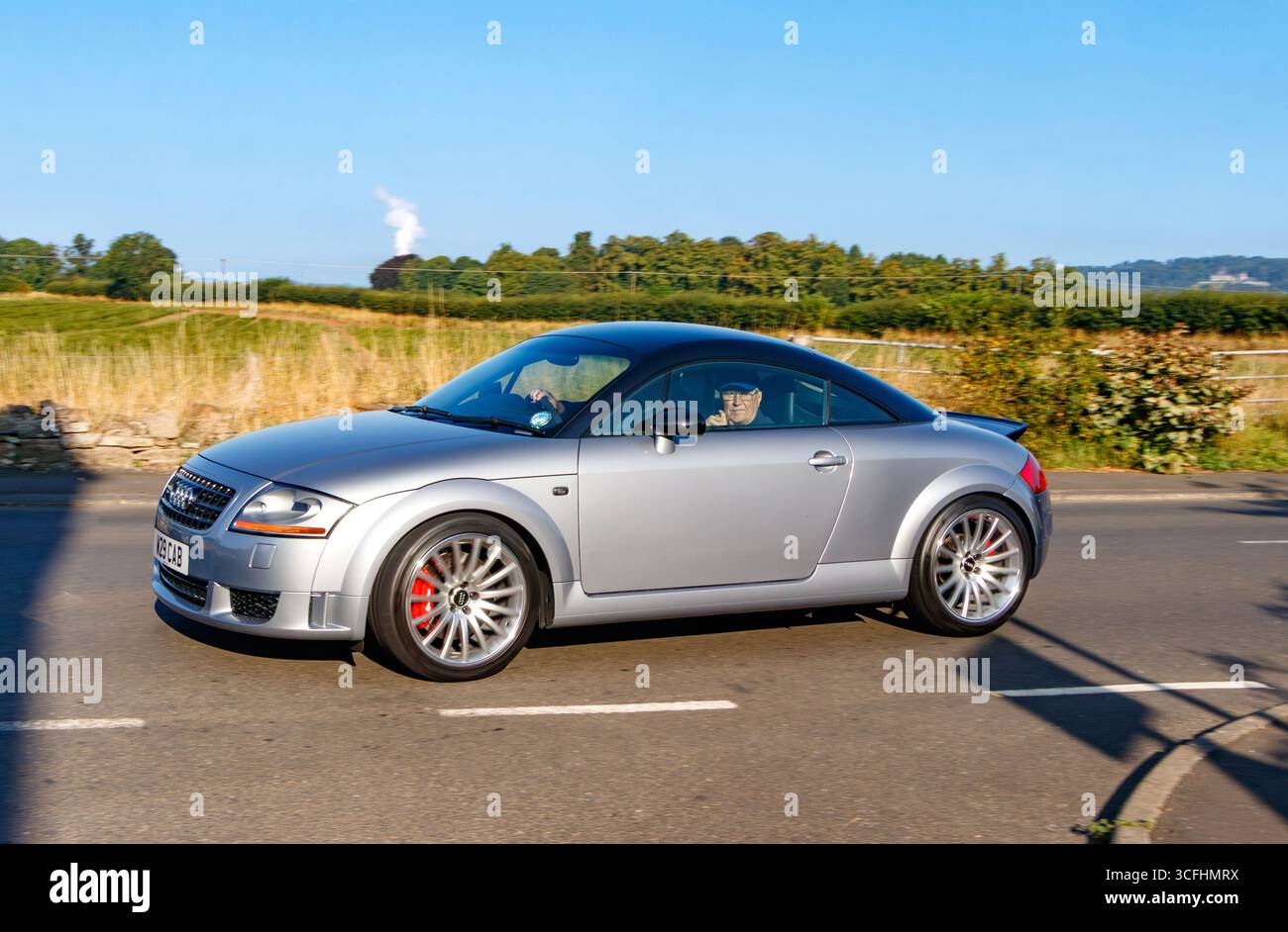 Auto sportiva d'argento che guida su una strada rurale sotto un cielo azzurro, con campi e alberi sullo sfondo in una giornata di sole Foto Stock