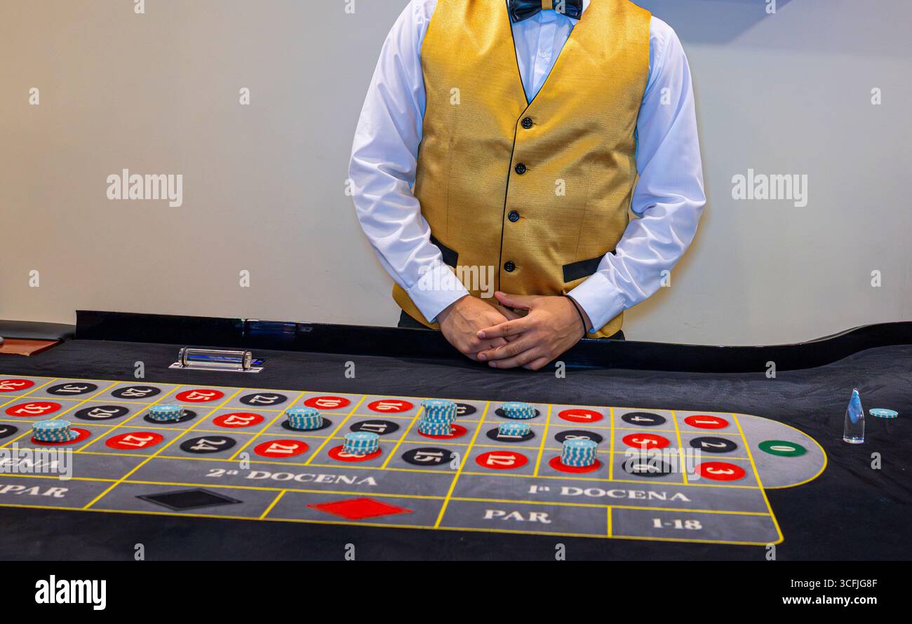 Elegante croupier in gilet dorato in piedi al tavolo della roulette con patatine pronte per giocare al casinò e per giocare d'azzardo. Foto Stock