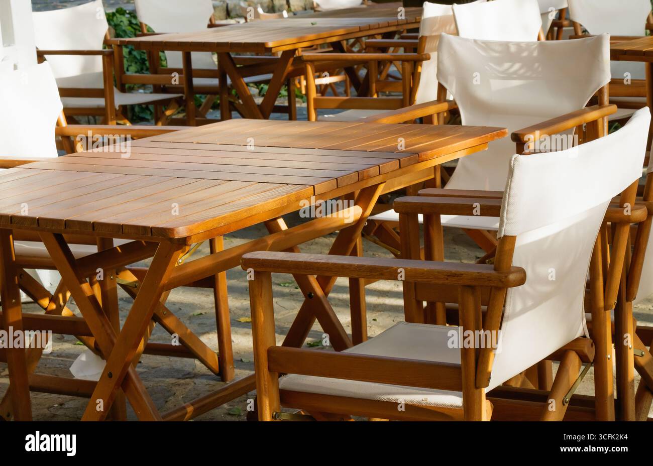 Un'accogliente area salotto all'aperto con tavoli in legno e sedie in tessuto bianco, allestita in un tranquillo giardino, illuminata da una delicata mattinata Foto Stock