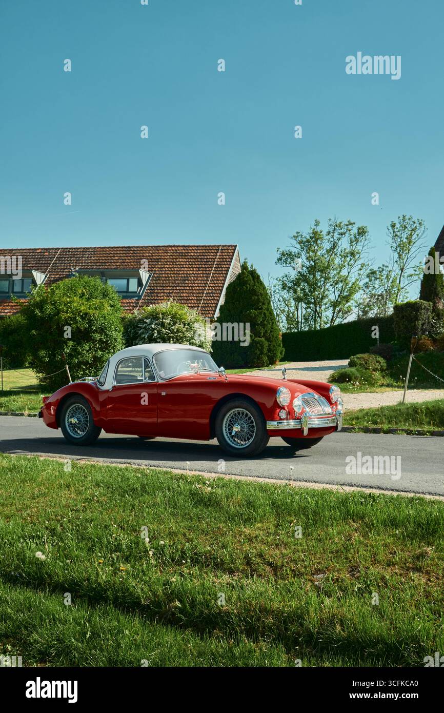Auto rossa retrò che guida su una strada con erba ben rifilata in primo piano, un edificio con un tetto rosso sullo sfondo e un cielo azzurro Foto Stock