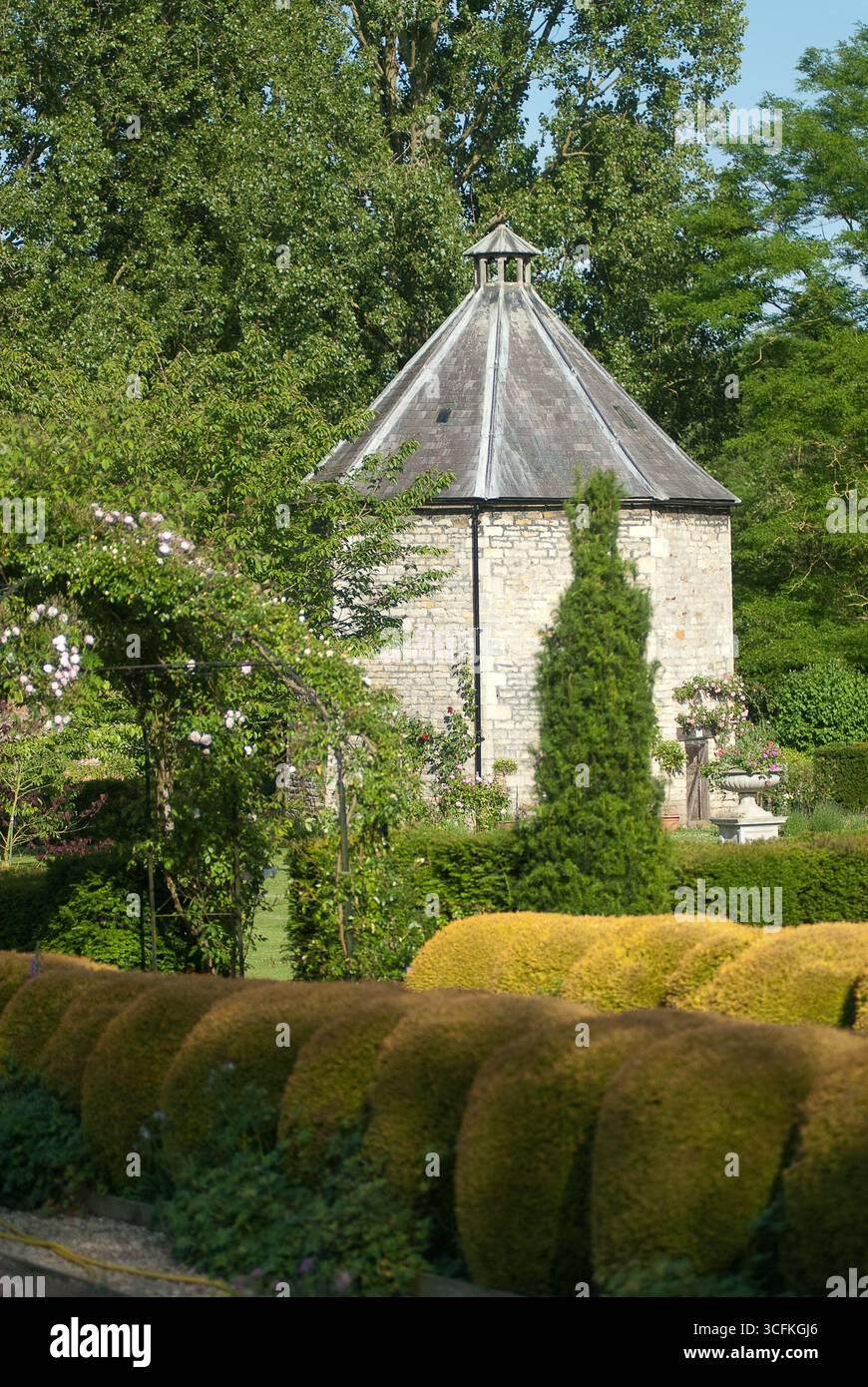 Un'affascinante struttura in pietra con un tetto unico sorge tra siepi vibranti e giardini ben tenuti, offrendo un tranquillo rifugio nella natura. Foto Stock