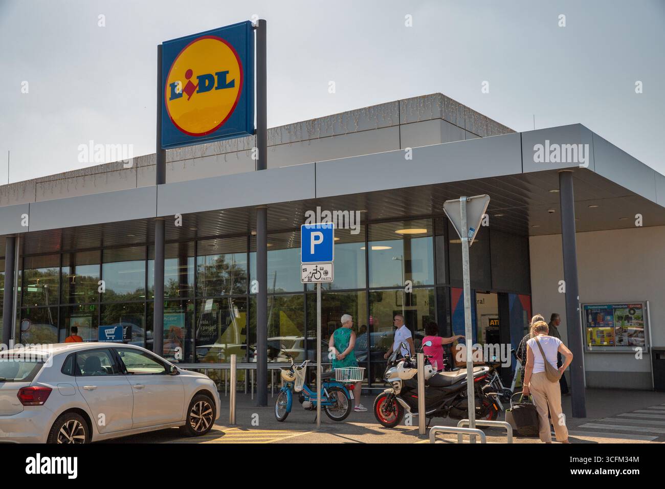 Porec, Croazia - 31 maggio 2025: Facciata moderna del negozio di alimentari Lidl con distintivo logo blu e giallo. Gli acquirenti entrano nel negozio con auto parcheggiate e bicicentro Foto Stock