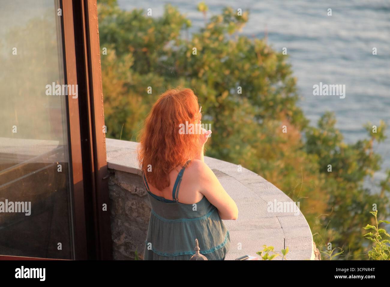 Donna capelli rossi che fuma e guarda dal balcone rotondo sul mare al tramonto, Ulcinj Stari Grad, Montenegro Foto Stock