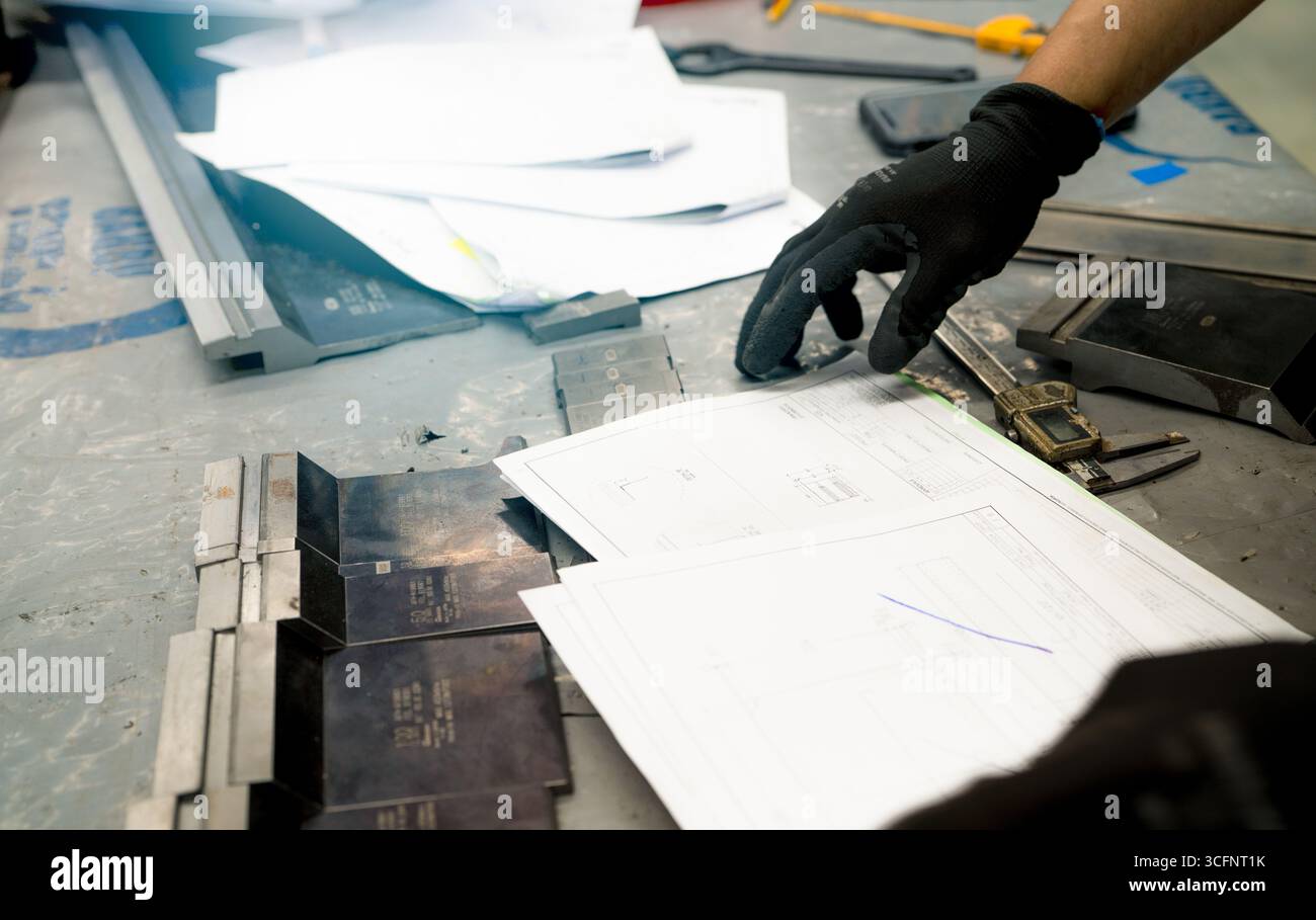 Mano del lavoratore con guanti neri che esaminano i disegni tecnici e gli attrezzi metallici su un tavolo da officina, evidenziando la precisione e le competenze coinvolte nell'industria dell'industria dell'induismo Foto Stock