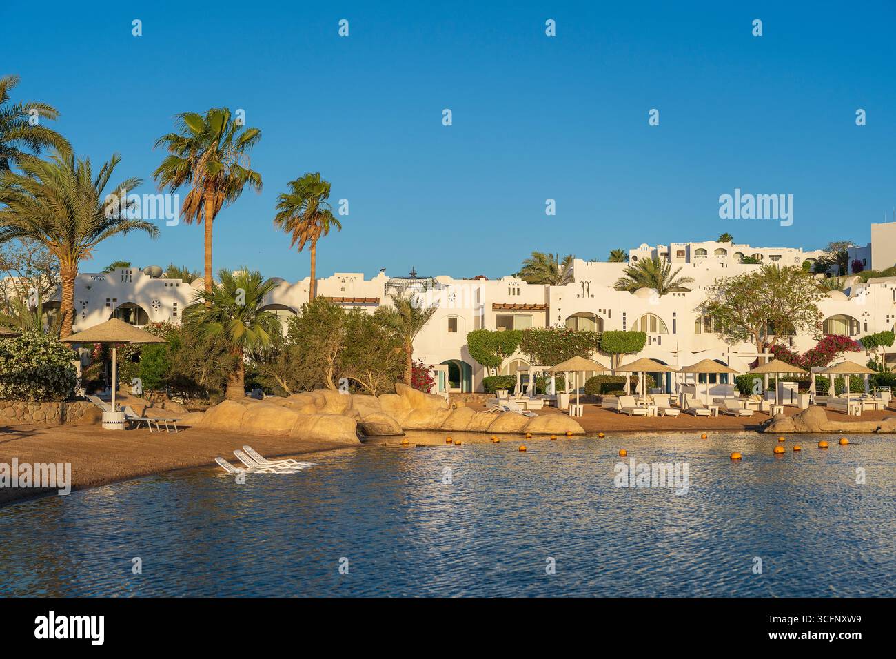 Edifici, sedie a sdraio e ombrelloni si riflettono nella calma acqua di mare sulla spiaggia nella località turistica di Sharm El Sheikh durante l'alba, in Egitto Foto Stock