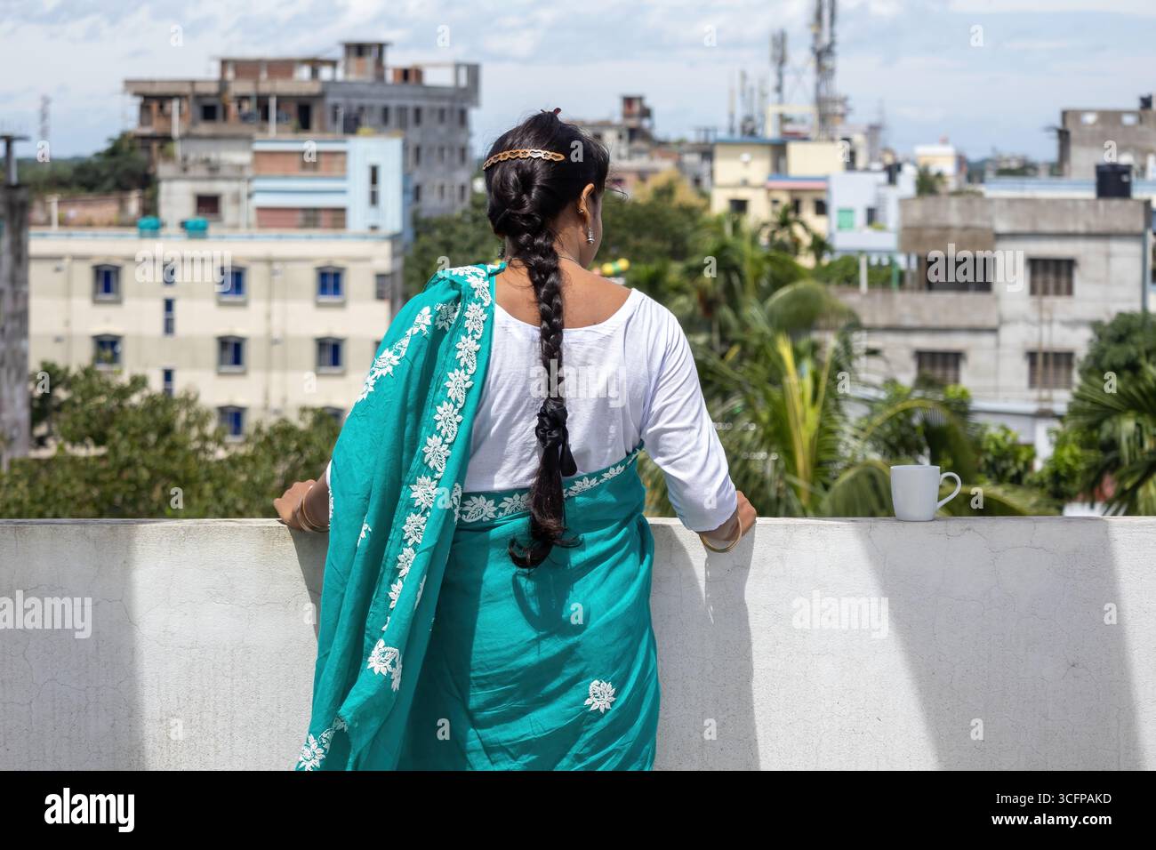 Donna bengalese in un sari turchese si erge su un tetto, i suoi lunghi capelli intrecciati cadono giù per la schiena. Guarda la città con una tazza di caffè accanto Foto Stock