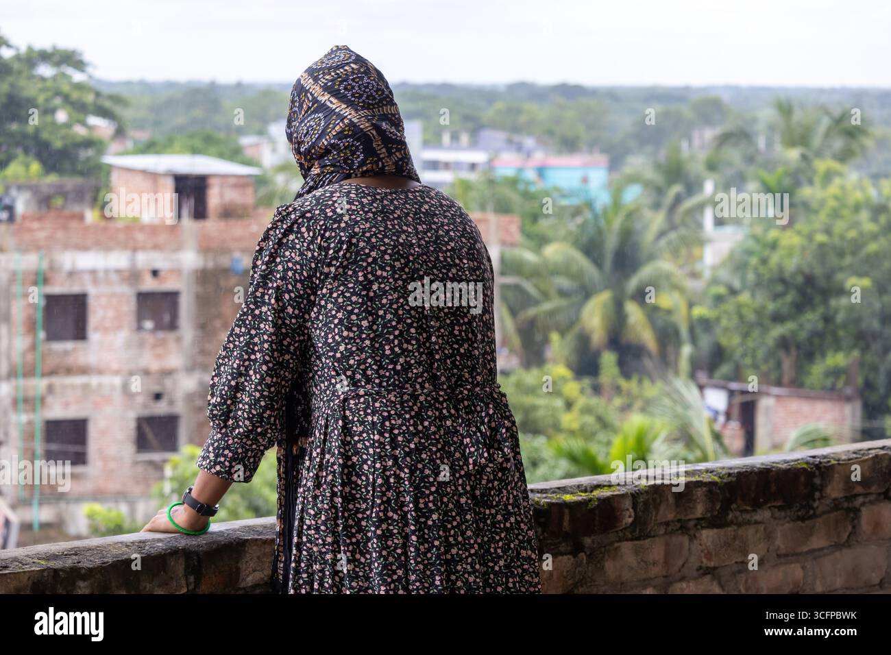 Vista posteriore di una premurosa donna musulmana bengalese in un abaya e hijab floreale, che guarda pacificamente la natura panoramica da un tetto. Foto Stock