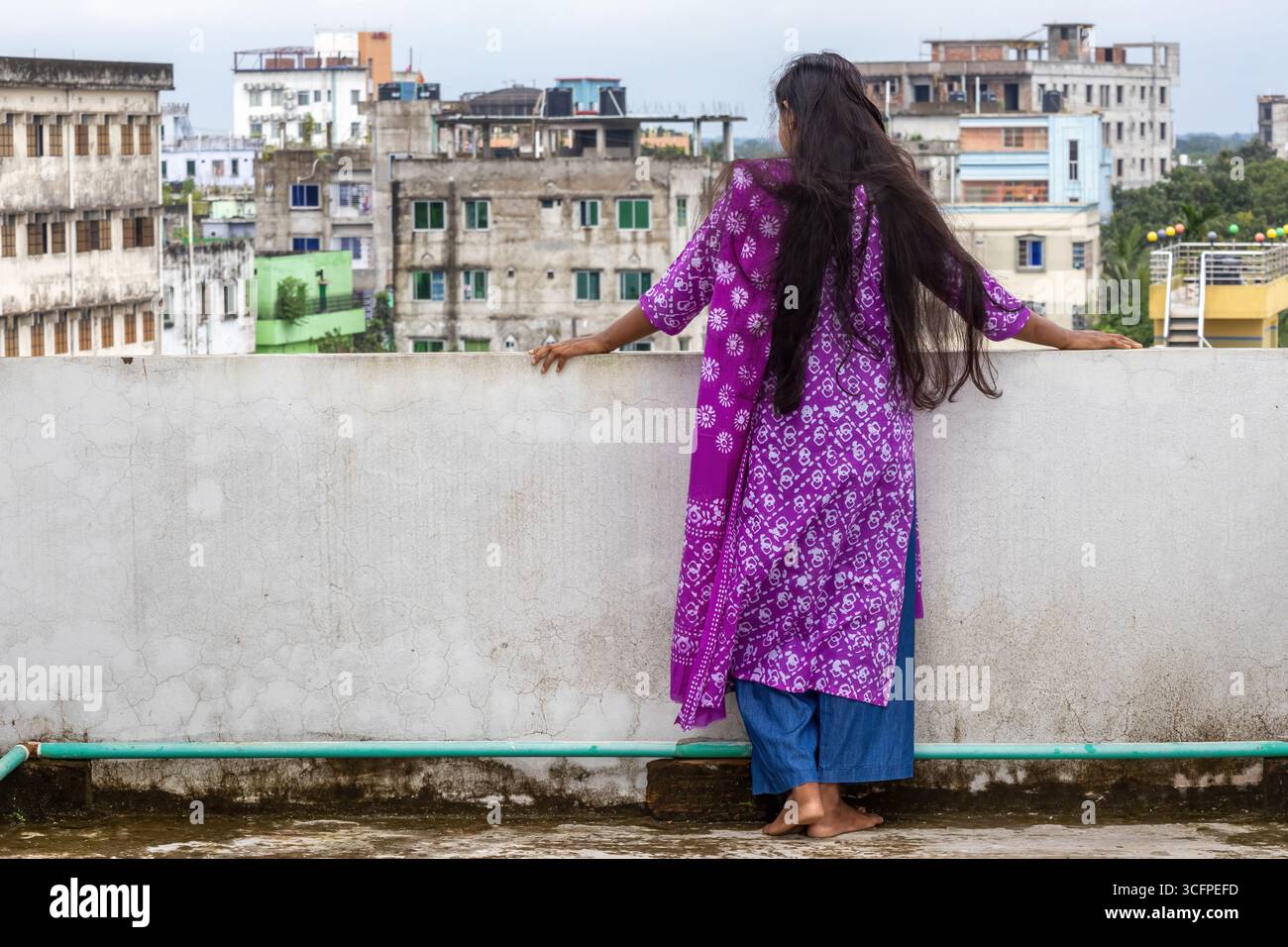 Splendida donna bengalese con lunghi capelli neri in un salwar kameez di cotone con stampa batik viola, con vista sulla città. E' da sola su una roo Foto Stock