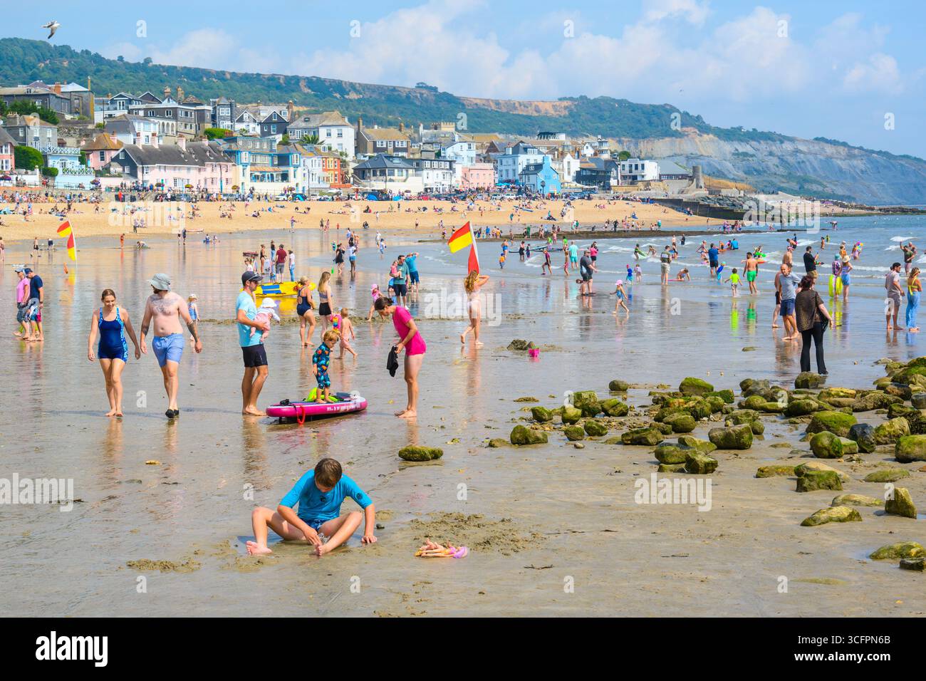 Lyme Regis, Dorset, Regno Unito. 24 agosto 2025. Meteo nel Regno Unito: Turisti, famiglie e amanti del sole pagaiano nel mare presso la località balneare di Lyme regis in un pomeriggio di sole rovente. Il lunedì festivo le temperature sono impostate per salire fino a 30 °C, con un bel tempo caldo e soleggiato da godersi prima che il clima umido e ventoso si diffonda in seguito all'uragano Erin. Crediti: Celia McMahon/Alamy Live News Foto Stock