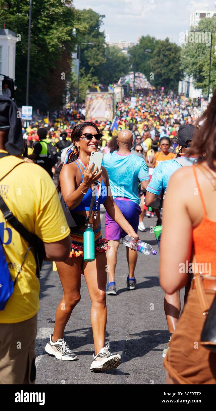 Folla al Carnevale di Notting Hill mentre una donna sorridente con gli occhiali da sole scatta foto al telefono e porta una bottiglia d'acqua a Londra. Inghilterra 24 agosto 2025 Foto Stock