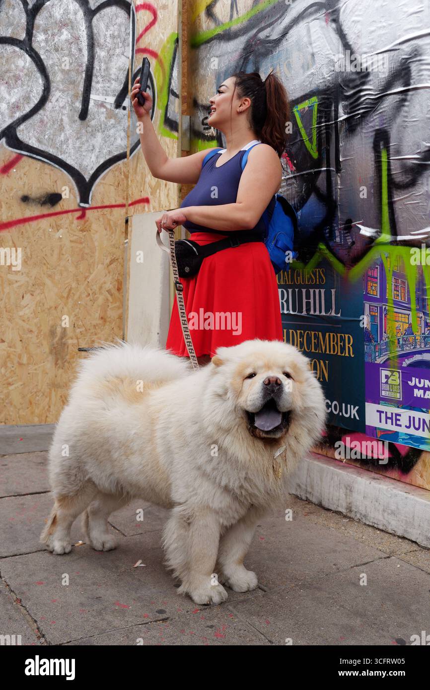 Una donna con un cane peloso molto grande si fa un selfie durante il Carnevale di Notting Hill, Londra. Inghilterra 24 agosto 2025 Foto Stock