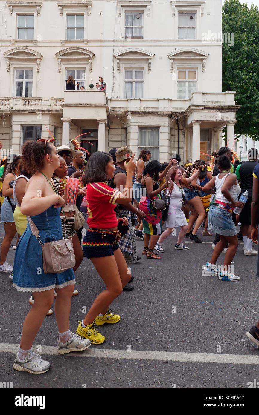 Gli spettatori ballano per strada con le proprietà dietro le quali si cela uno spirito di comunità durante il Carnevale di Notting Hill, Londra. Inghilterra 24 agosto 2025 Foto Stock