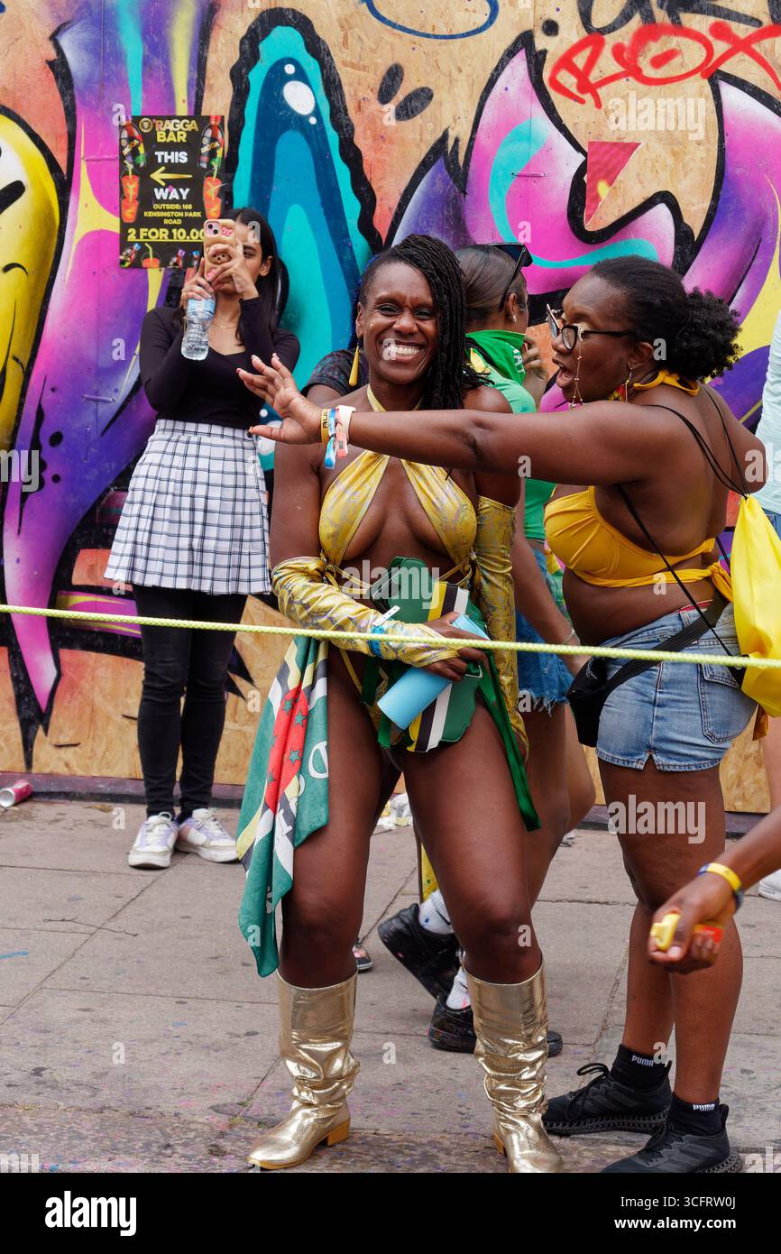 Donna nera in costume da donna meraviglia sorride ampiamente accanto alla sua amica durante il Carnevale di Notting Hill, Londra. Inghilterra 24 agosto 2025 Foto Stock