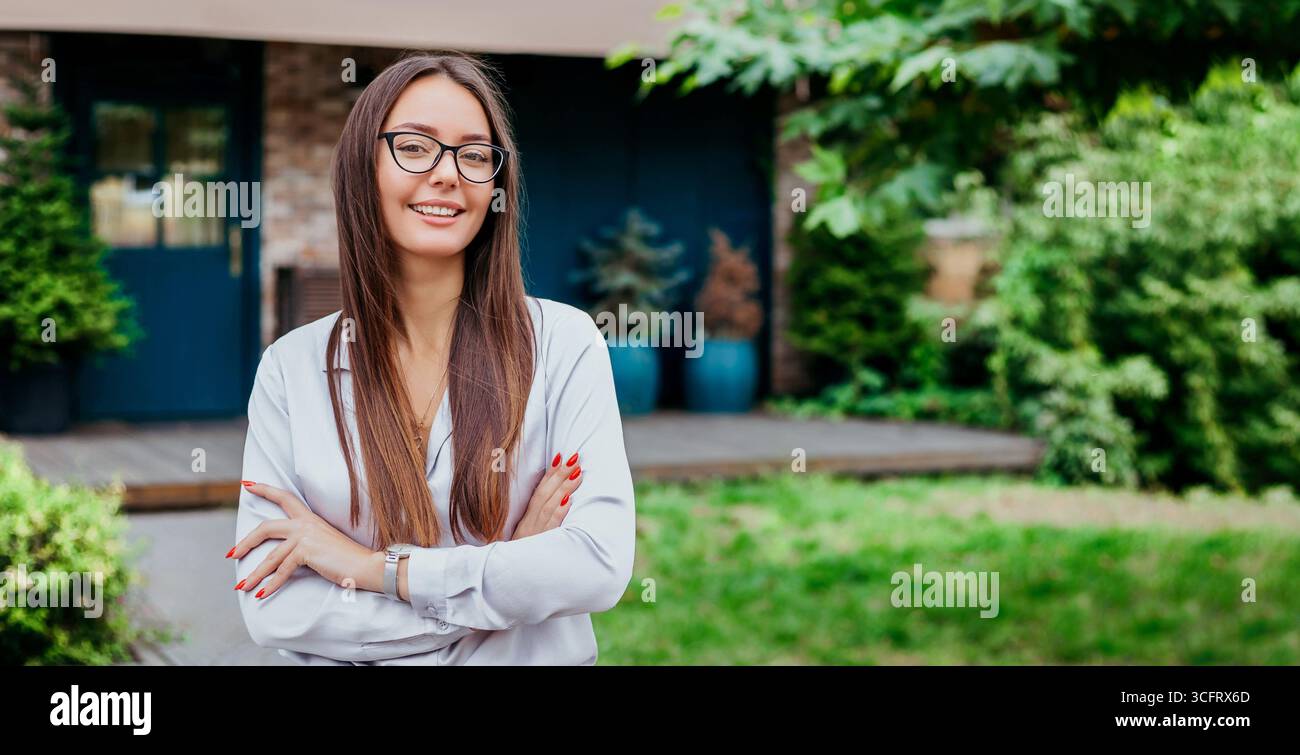 Una giovane studentessa in occhiali sorride con le braccia piegate vestita con una camicia sullo sfondo della facciata dell'edificio, copia spazio Foto Stock