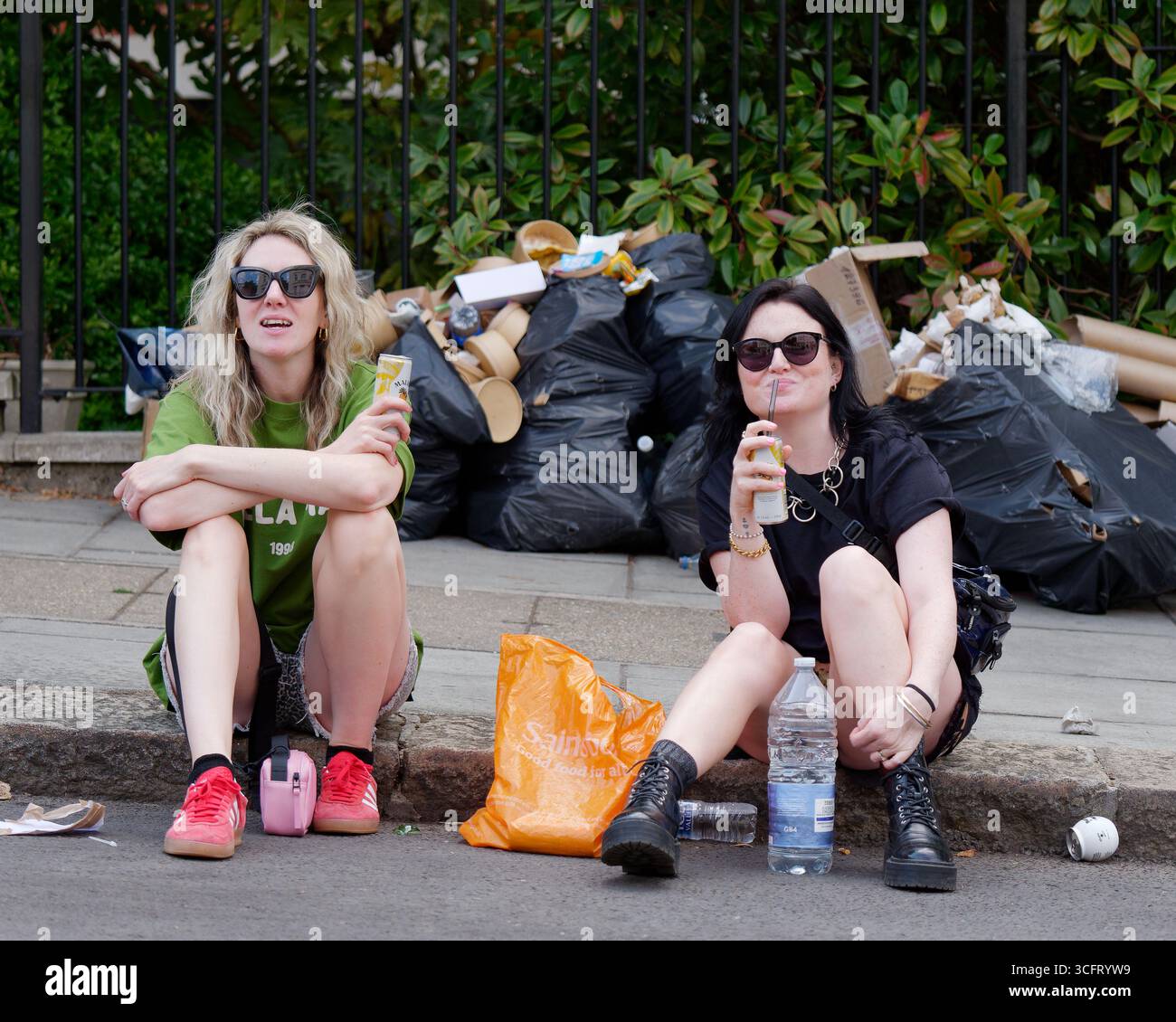 Due spettatori femminili siedono sul marciapiede con i rifiuti dietro durante il Carnevale di Notting Hill, Londra. Inghilterra 25 agosto 2025 Foto Stock