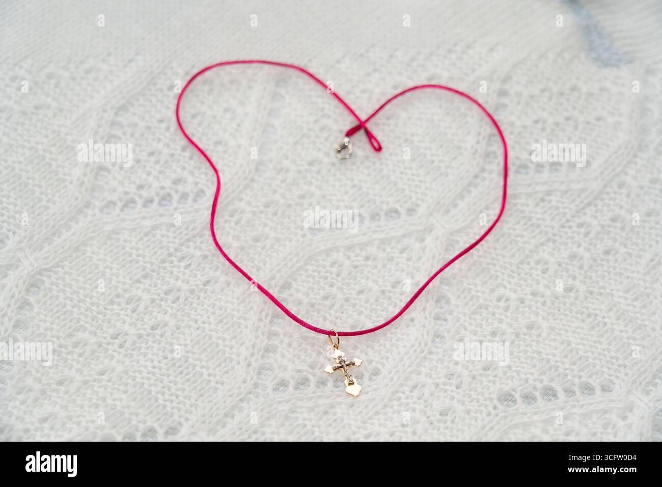 Una collana rosa con cordoncino e un piccolo ciondolo a croce è disposta a forma di cuore su un bianco. Il design trasuda semplicità ed eleganza, ideale per ogni occasione Foto Stock