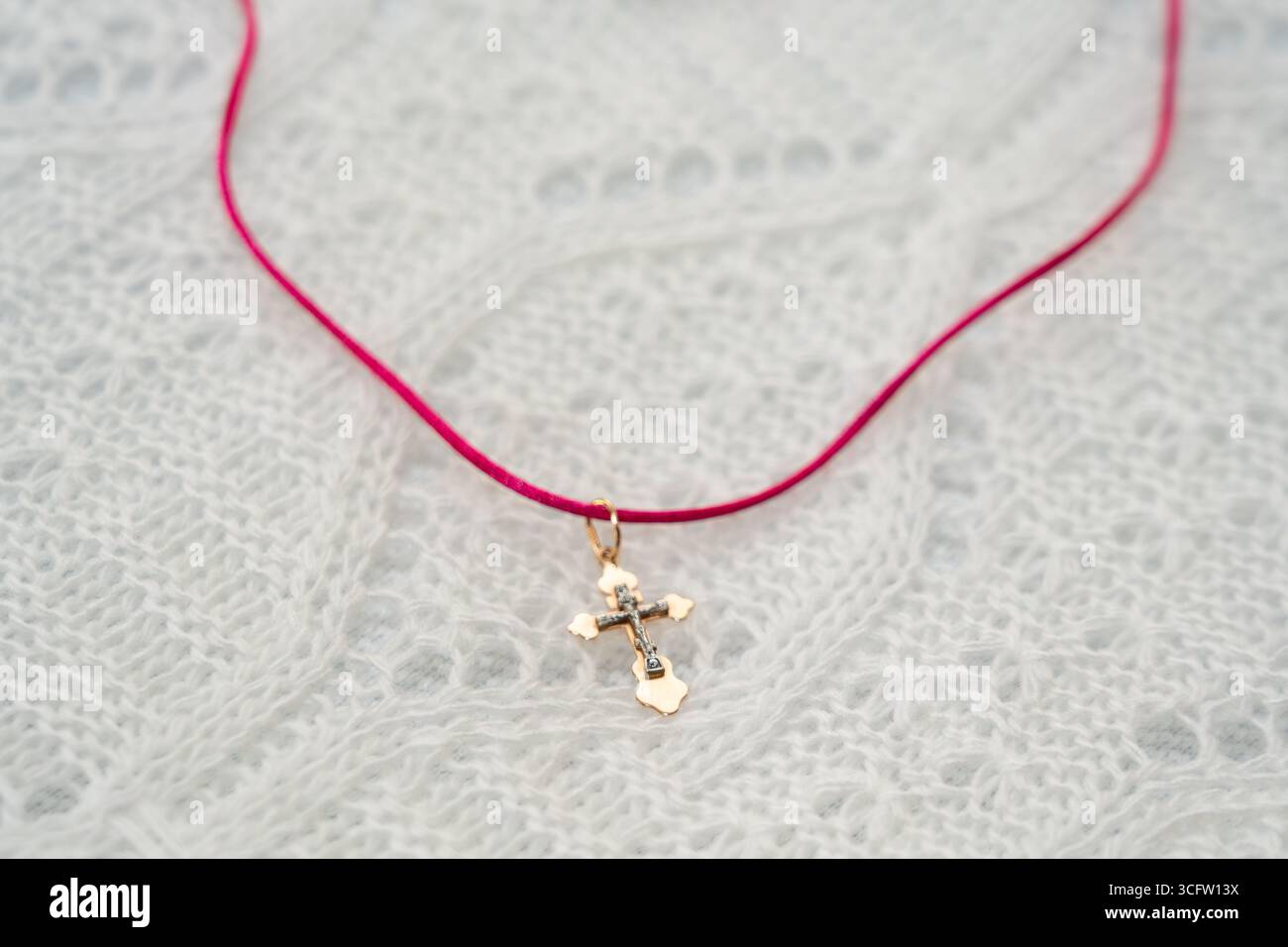 Un delicato ciondolo a croce dorato è appeso a un filo rosso, appoggiato su un tessuto bianco testurizzato. Foto Stock