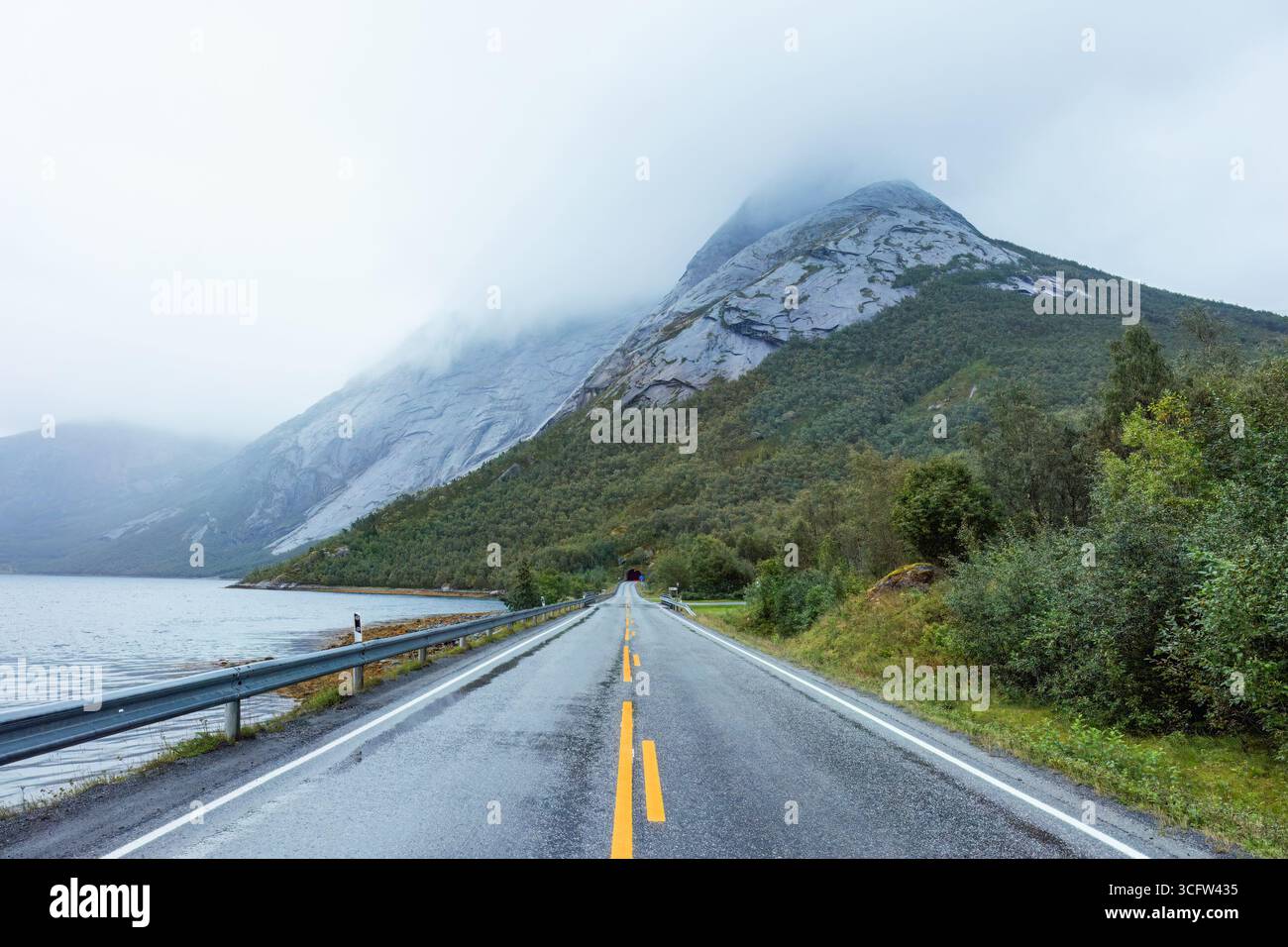 La strada tra le montagne e il lago, immersa nella nebbia, offre una tranquilla fuga nella bellezza della natura nordica. Stetind Mountain, Nordland, Norvegia Foto Stock