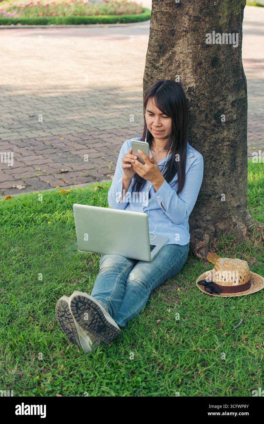 Donna asiatica seduta verde parco giardino utilizzando il telefono ascoltare la musica da smartphone con computer portatile. Felicità donna sorridente, ridendo felice toothy s Foto Stock
