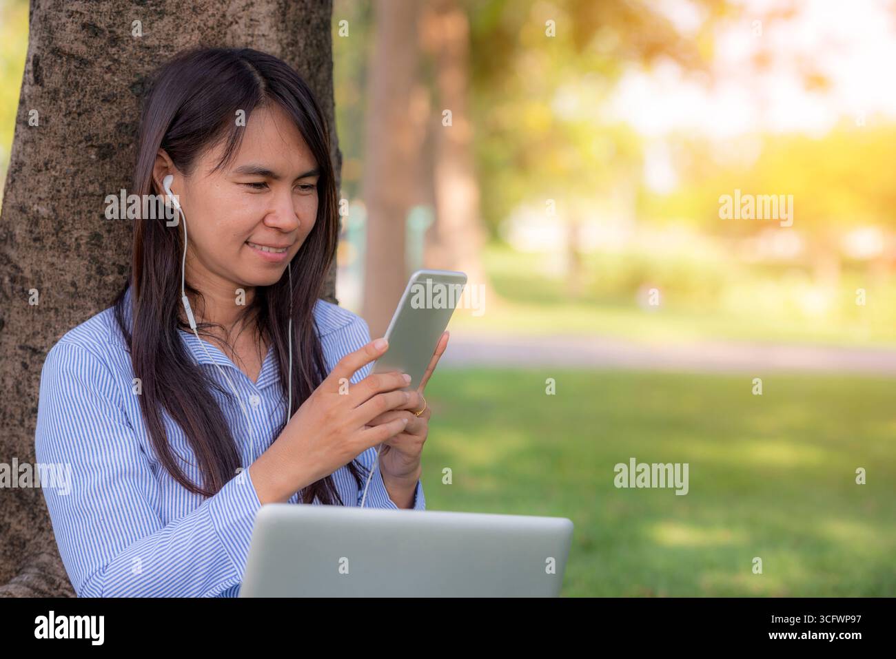 Donna asiatica seduta verde parco giardino utilizzando il telefono ascoltare la musica da smartphone con computer portatile. Felicità donna sorridente, ridendo felice toothy s Foto Stock
