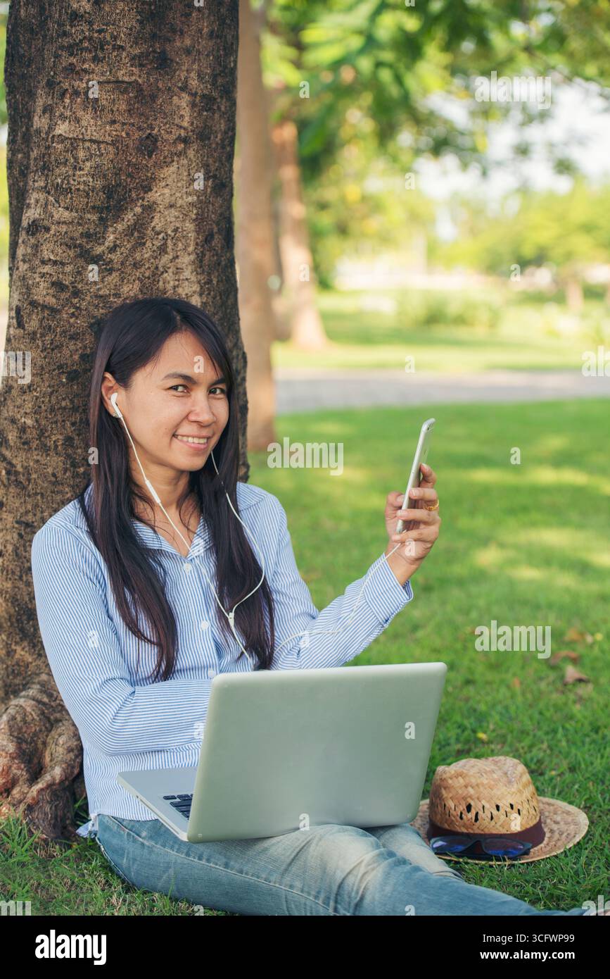 Donna asiatica seduta verde parco giardino utilizzando il telefono ascoltare la musica da smartphone con computer portatile. Felicità donna sorridente, ridendo felice toothy s Foto Stock