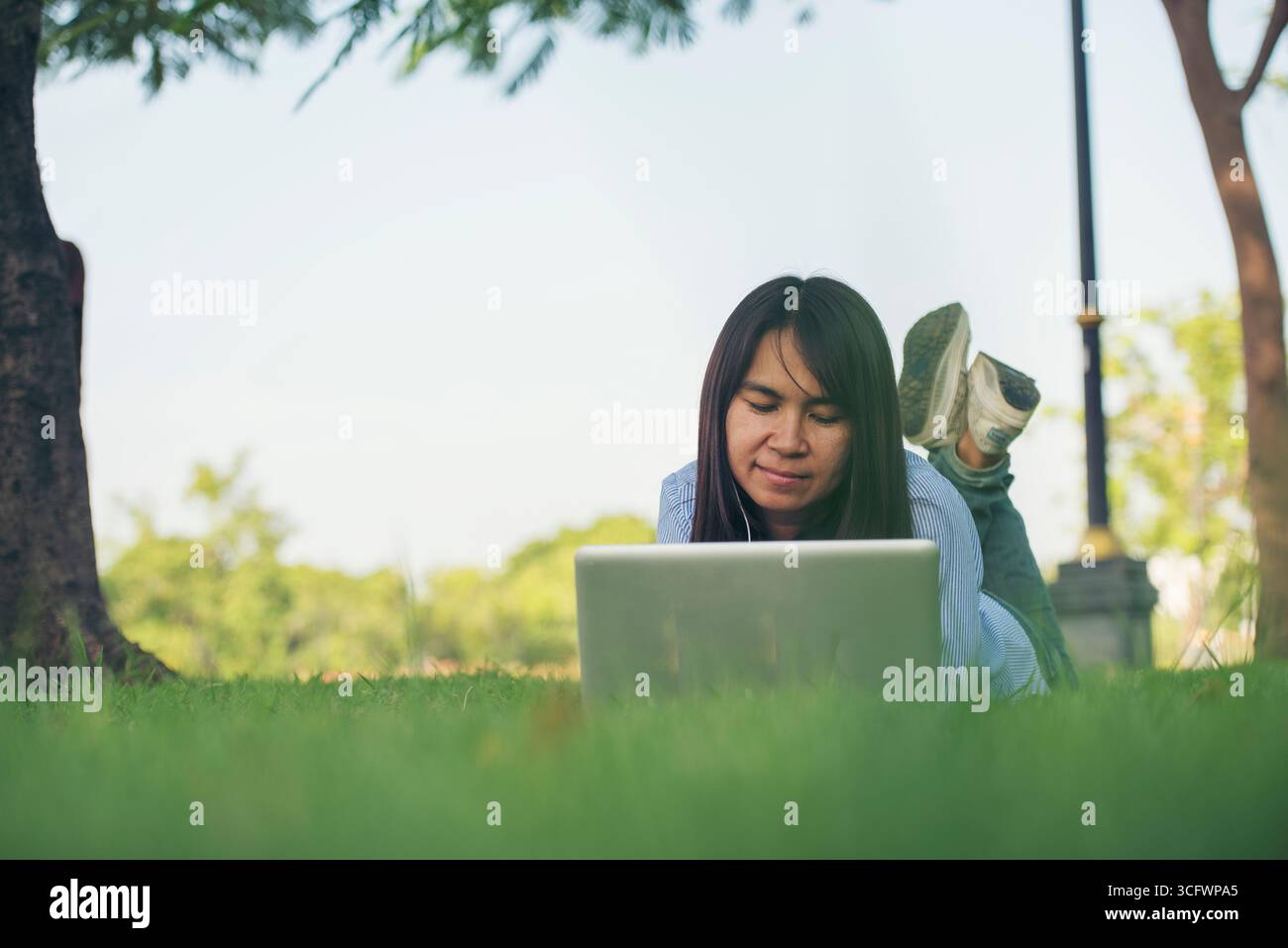 Donna asiatica stendersi parco verde giardino utilizzando computer portatile che lavora all'aperto casa ufficio. La felicità della donna sorride lavorando freelance su un notebook. Giovani donne Univer Foto Stock