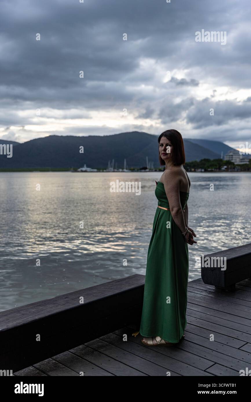 lato su una donna occhi che ti guardano con un abito verde che posa in piedi su una passerella di legno una donna elegante che indossa un abito verde contro una scenografica spiaggia Foto Stock lato su una donna occhi che ti guardano con un abito verde che posa in piedi su una passerella di legno una donna elegante che indossa un abito verde contro una scenografica spiaggia Foto Stock