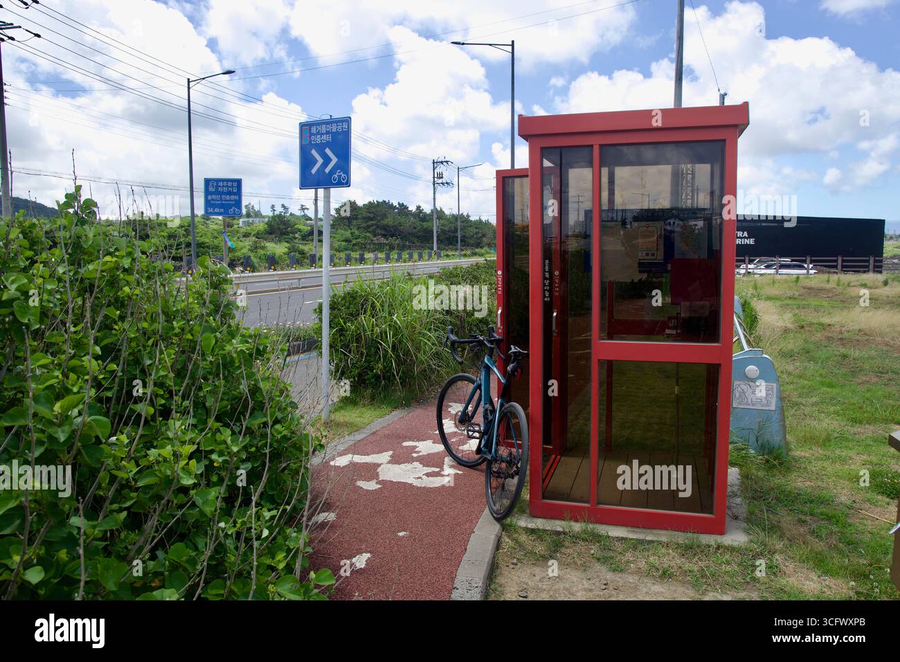 Una bicicletta si appoggia sul rifugio rosso del Parco Haegeoreum, accanto a un cartello blu di certificazione del percorso ciclabile Jeju circolare lungo l'autostrada costiera. Foto Stock