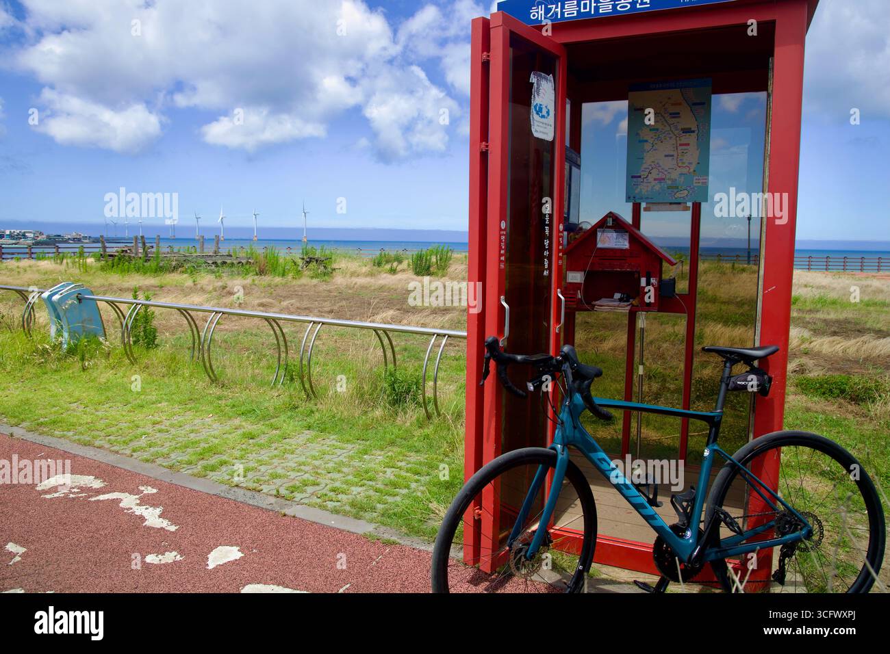 Un rifugio certificato per ciclismo rosso al parco Haegeoreum si trova accanto a un portabiciclette e all'erba costiera, con una bici da strada appoggiata contro di essa e vento offshore Foto Stock