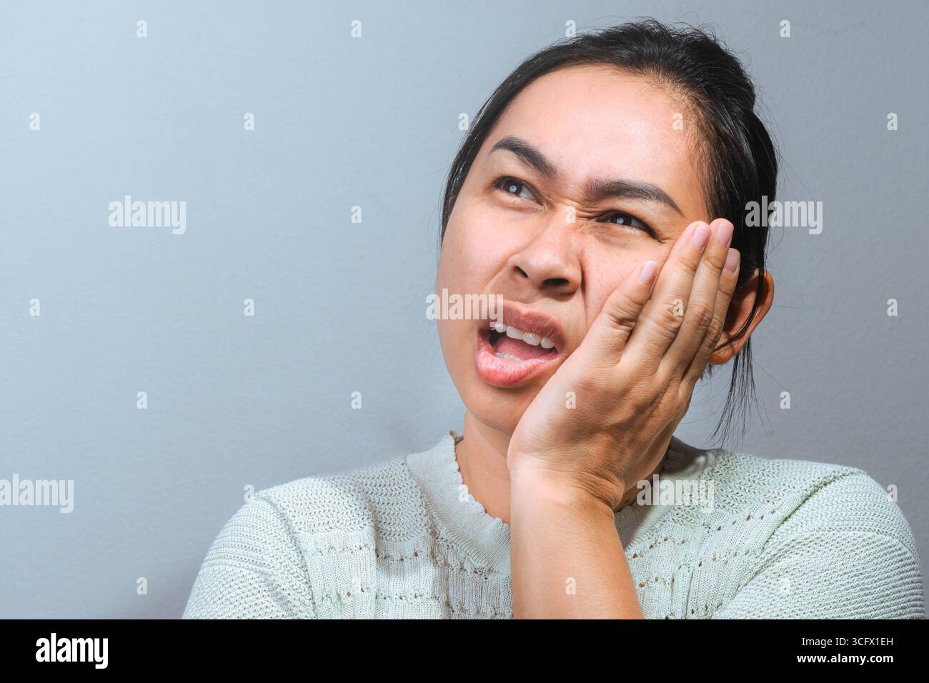 Una donna asiatica che tiene la guancia con un'espressione dolorosa, mostrando disagio come se soffrisse di mal di denti o problemi dentali. Foto Stock