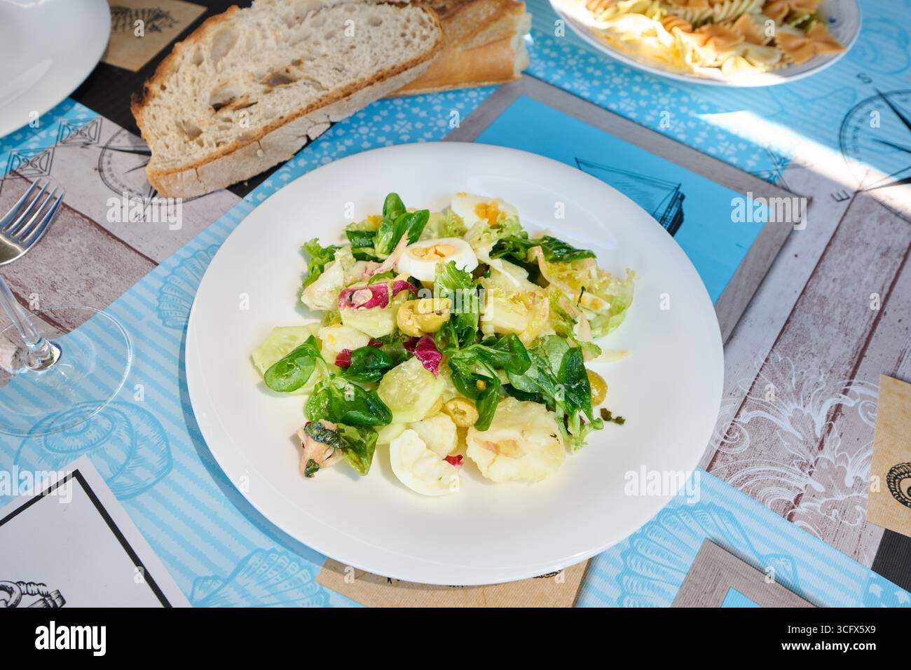 Vivace insalata mista con uova sode, cetrioli, olive e lattuga, servita su un piatto con tovaglietta a tema nautico, accompagnata da pane e pasta Foto Stock
