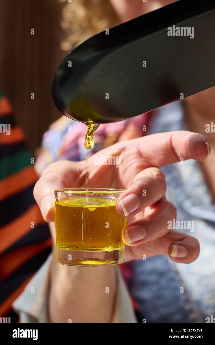 Primo piano della mano di una donna che tiene un piccolo bicchiere mentre versa l'olio d'oliva da una paletta di metallo, durante una degustazione di olio d'oliva Foto Stock