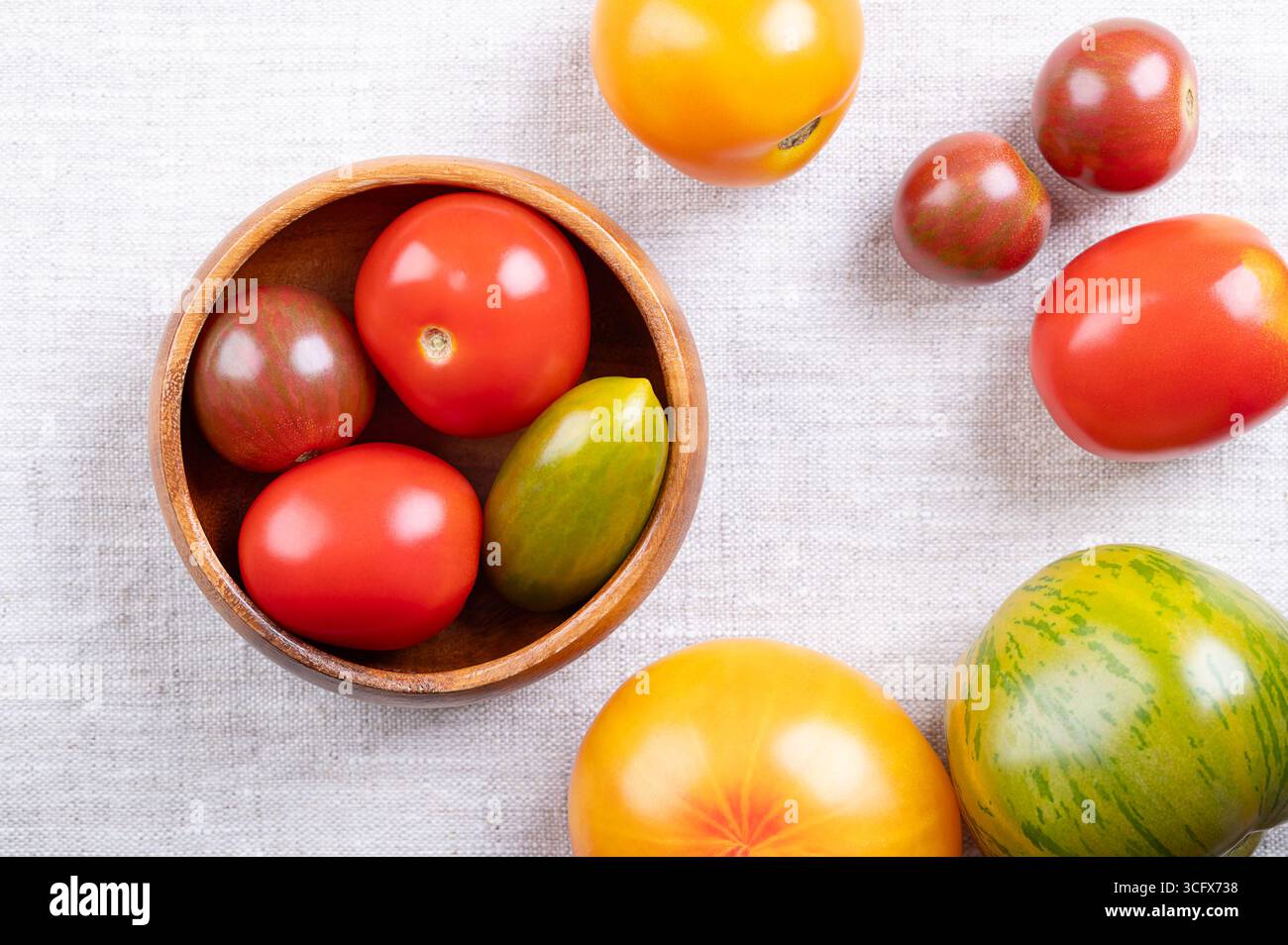 Pomodori freschi e maturi di varie varietà, colori e dimensioni in una ciotola di legno, su lino. Varietà della pianta Solanum lycopersicum. Foto Stock