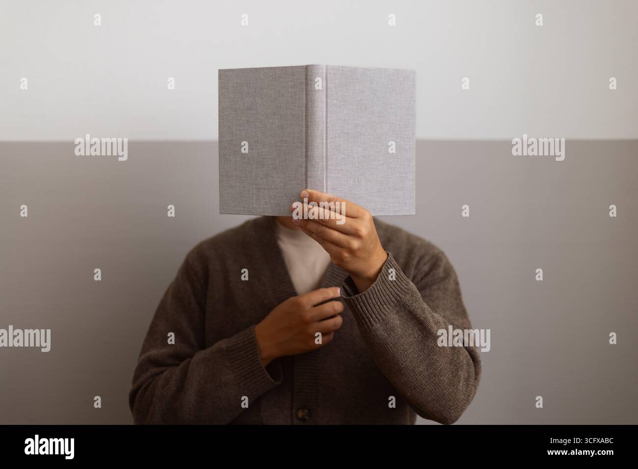 Persona che tiene un libro aperto davanti al viso, creando una scena di lettura minimale e tranquilla. Toni neutri e sfondo semplice. Foto Stock