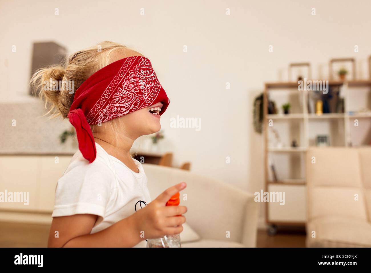 Un bambino che indossa una bandana rossa bendata e tiene in mano una bottiglia spray, sorridendo gioiosamente in un moderno soggiorno luminoso, impegnandosi in un giocoso a Foto Stock