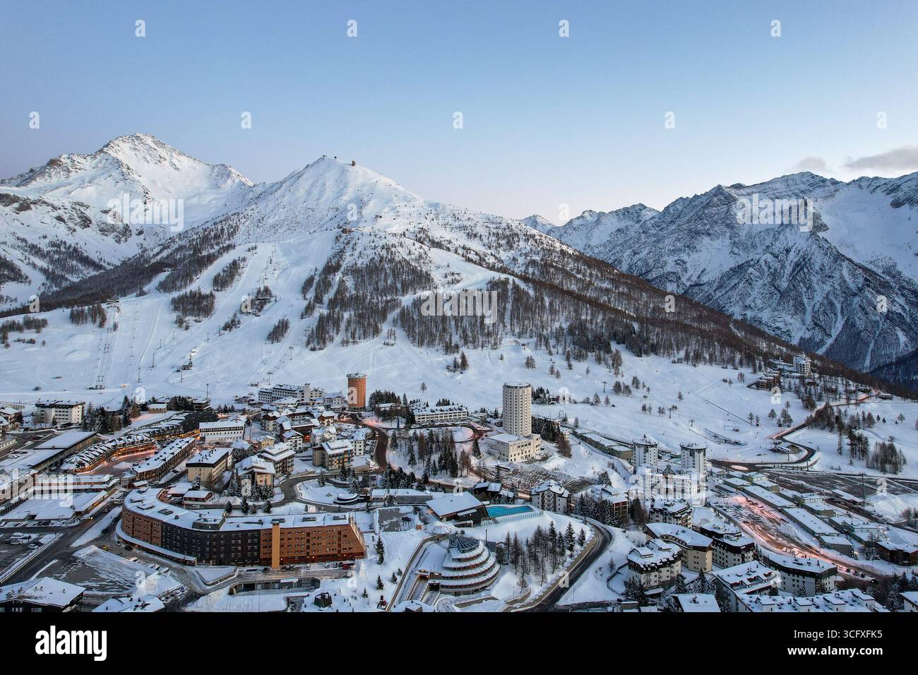 Veduta aerea di edifici innevati annidati tra torreggianti montagne innevate contro un cielo pallido, Sestriere, Piemonte, Italia. Foto Stock