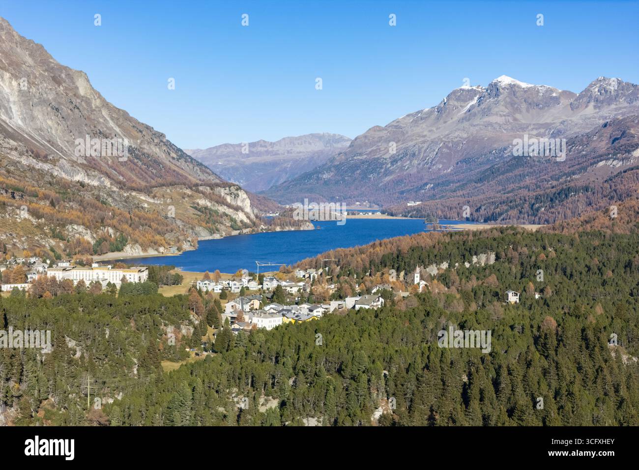 Vista aerea del tranquillo lago Silsersee che riflette il cielo azzurro, annidato tra le aspre montagne dai toni autunnali e le lussureggianti foreste, Maloja Foto Stock