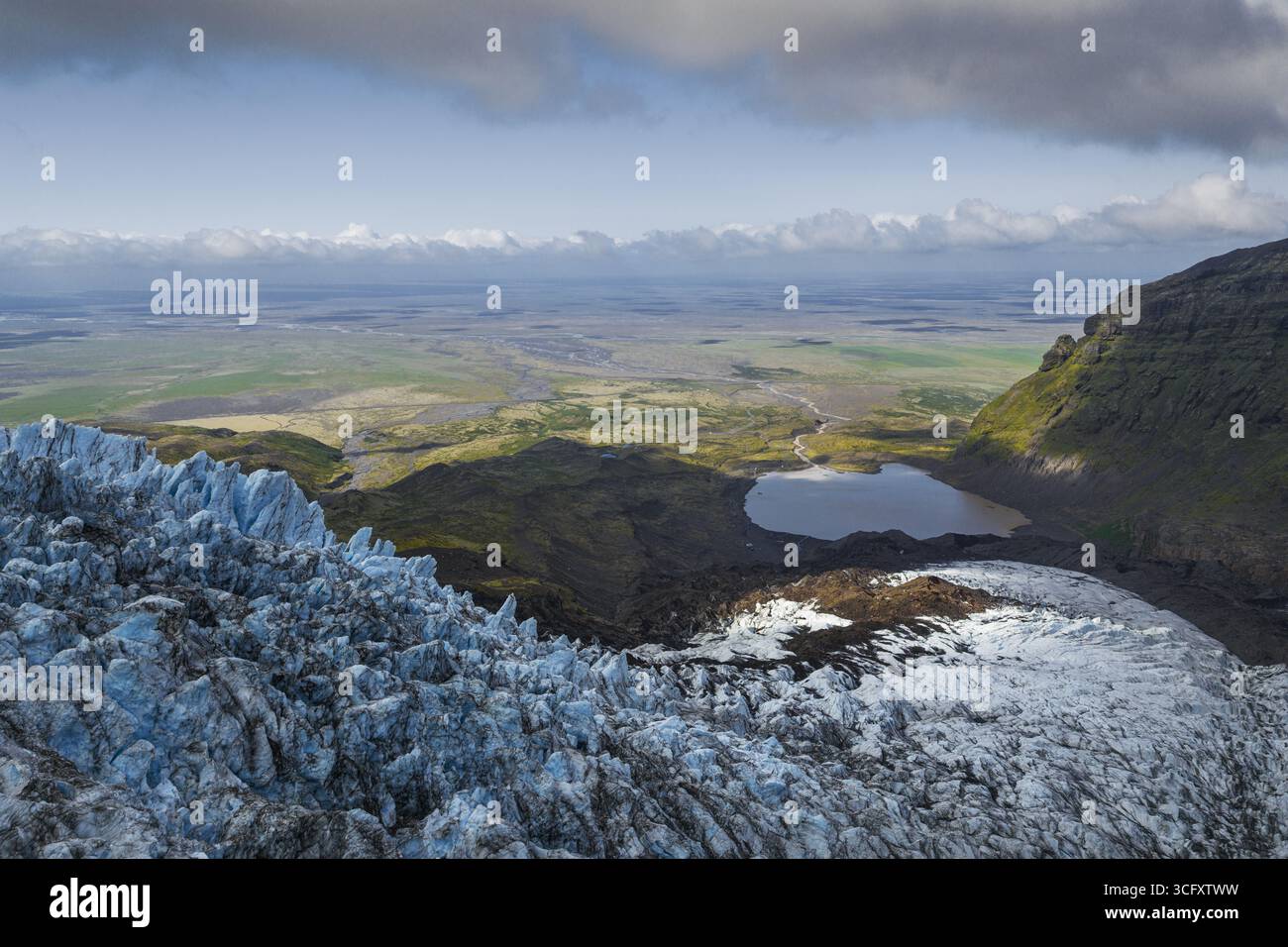 La vista aerea delle sfumature blu ghiacciate del ghiacciaio incontra il paesaggio aspro e scuro, in contrasto con le lontane pianure verdi sotto un cielo di morbide nuvole, altopiani e Islanda. Foto Stock