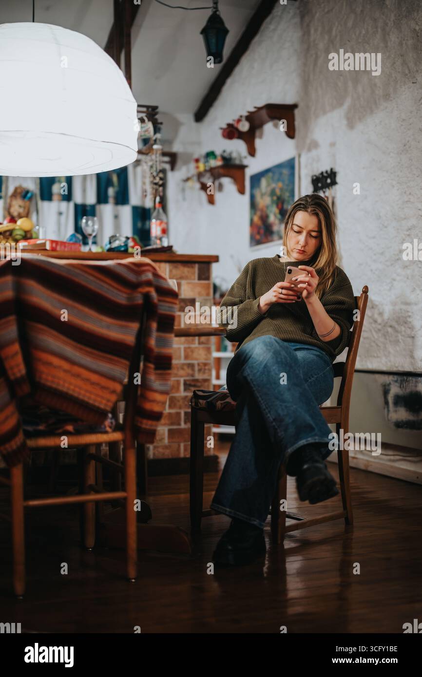 Giovane donna seduta in un'accogliente cucina che controlla il suo smartphone con attenzione e coinvolgimento Foto Stock
